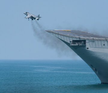 A military jet performing a short takeoff using a ski-jump ramp from the bow of an amphibious assault ship or aircraft carrier.