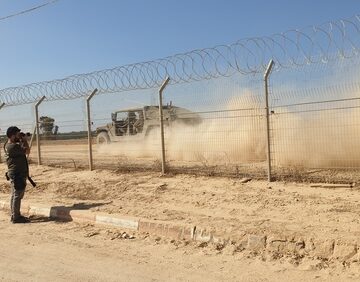 A security fence line at the Gaza border with barbed wire, a vehicle kicking up dust, and an individual observing the area. A security fence line at the Gaza border with barbed wire, a vehicle kicking up dust, and an individual observing the area.