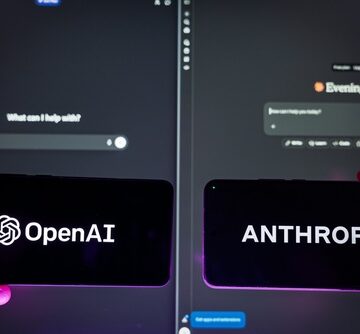 The image shows two phones displaying the logos of competing artificial intelligence companies, OpenAI and Anthropic, held up against computer monitors displaying respective AI interfaces.