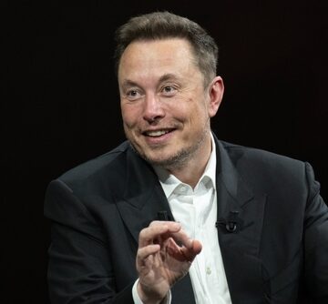 Entrepreneur and business leader Elon Musk gesturing while wearing a dark suit jacket over a white dress shirt.