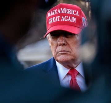 Donald Trump wearing a red "Make America Great Again" cap, a dark blue suit, and a red tie.