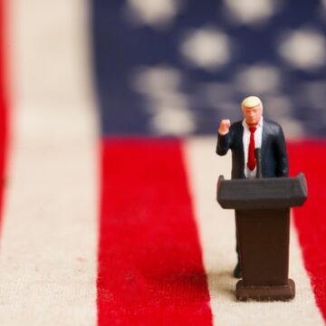 A miniature figurine of Donald Trump standing behind a lectern with his right fist raised, positioned in front of a United States flag background.