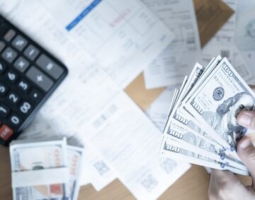 A person counting stacks of U.S. dollar bills next to a black desktop calculator and financial documents. A person counting stacks of U.S. dollar bills next to a black desktop calculator and financial documents.