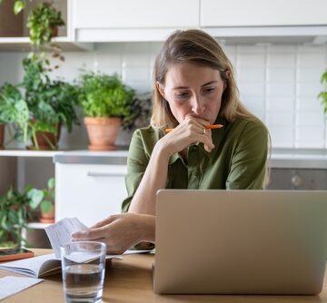 A concerned woman sits at a table with a laptop, reviewing household bills or paperwork. A concerned woman sits at a table with a laptop, reviewing household bills or paperwork.