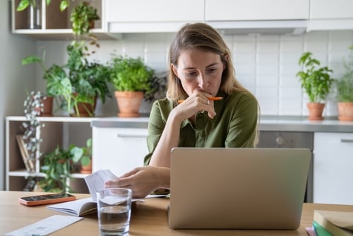A concerned woman sits at a table with a laptop, reviewing household bills or paperwork. A concerned woman sits at a table with a laptop, reviewing household bills or paperwork.