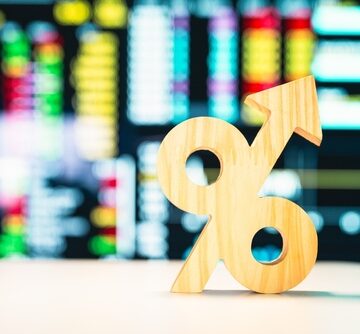 A wooden percentage symbol with an upward-pointing arrow in the foreground and blurred stock data on a screen is in the background. A wooden percentage symbol with an upward-pointing arrow in the foreground and blurred stock data on a screen is in the background.