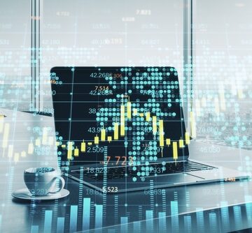 A laptop on an office desk with a digital financial candlestick chart and world map hologram overlay. A laptop on an office desk with a digital financial candlestick chart and world map hologram overlay.