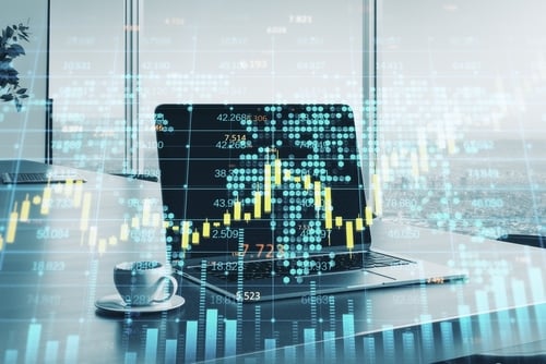 A laptop on an office desk with a digital financial candlestick chart and world map hologram overlay.