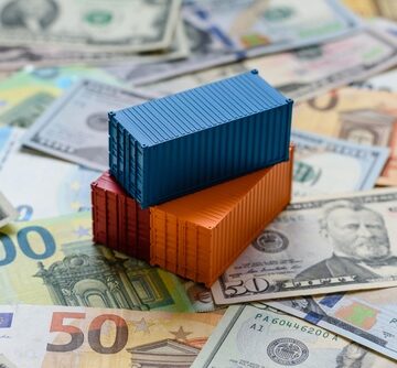 Miniature blue, orange, and red cargo shipping containers resting on top of a mix of U.S. Dollar and Euro banknotes, symbolizing international trade. Miniature blue, orange, and red cargo shipping containers resting on top of a mix of U.S. Dollar and Euro banknotes, symbolizing international trade.