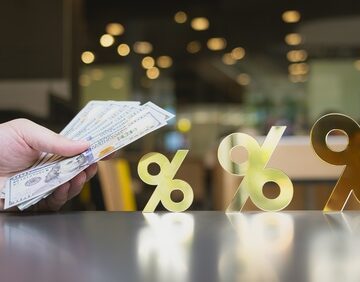 The image shows a hand holding U.S. dollar bills next to golden percentage signs on a table.