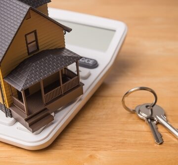 A small yellow house model resting on a white calculator next to a set of house keys on a wooden surface. A small yellow house model resting on a white calculator next to a set of house keys on a wooden surface.