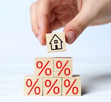 Five wooden blocks arranged in a pyramid shape, each marked with a red percent symbol (%) and the top wooden cube featuring a simple black outline drawing of a house being placed on top of the percentage pyramid by a person's fingers. Five wooden blocks arranged in a pyramid shape, each marked with a red percent symbol (%) and the top wooden cube featuring a simple black outline drawing of a house being placed on top of the percentage pyramid by a person's fingers.