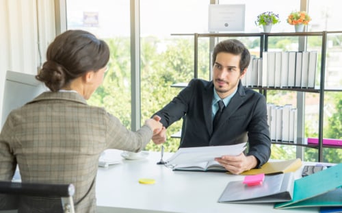 A professional business meeting or job interview setting where two people are shaking hands across a desk. A professional business meeting or job interview setting where two people are shaking hands across a desk.