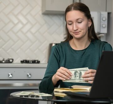 A woman in a green top sitting at a table in a kitchen, counting US dollar banknotes next to a laptop and a notebook.
