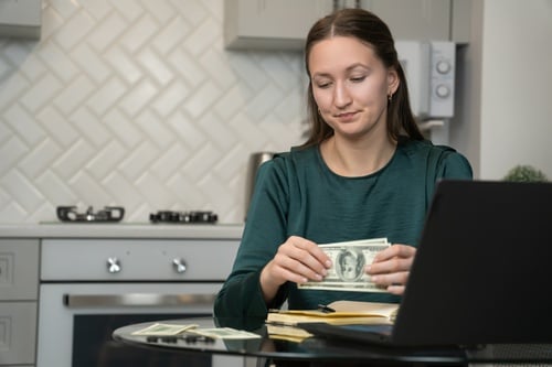 A woman in a green top sitting at a table in a kitchen, counting US dollar banknotes next to a laptop and a notebook. A woman in a green top sitting at a table in a kitchen, counting US dollar banknotes next to a laptop and a notebook.