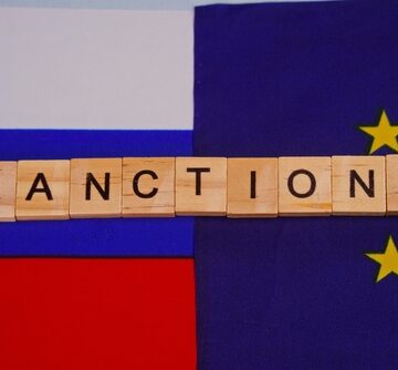 The word "SANCTIONS" spelled out on wooden tiles, positioned across the flags of Russia and the European Union.