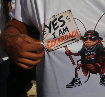 A white t-shirt featuring a graphic of an anthropomorphic cockroach wearing sunglasses and sneakers, holding a sign that says "YES I AM COCKROACH" and wearing a shirt reading "NOT A BUG, A VIBE".