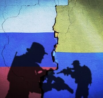 Conceptual artwork depicting two armed soldiers casting shadows against a cracked wall split between the flags of Russia and Ukraine.